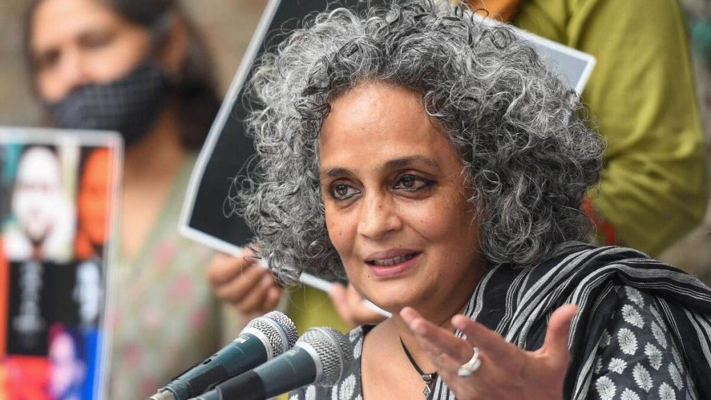 Arundhati Roy, autora india, hablando en un micrófono durante un evento público con audiencia de fondo