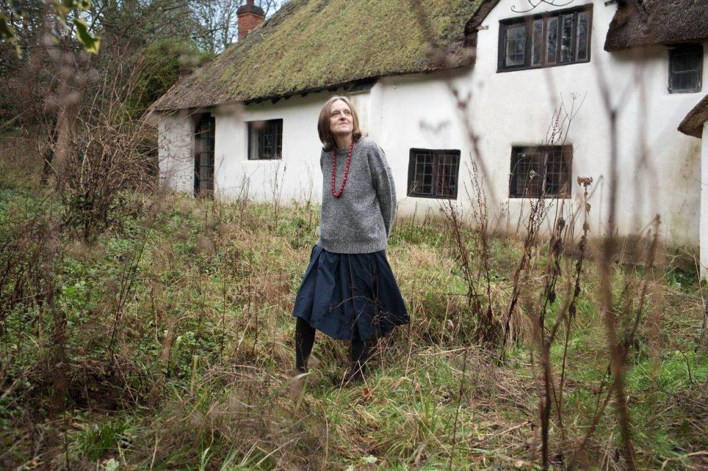 Tessa Hadley de pie frente a una casa blanca con tejado de paja, rodeada de vegetación salvaje y árboles