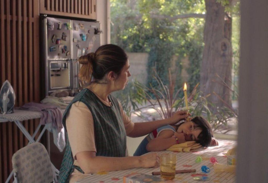 Mujer en interior luminoso sostiene a niña pequeña junto a ventana con vistas a naturaleza, escena cotidiana y íntima