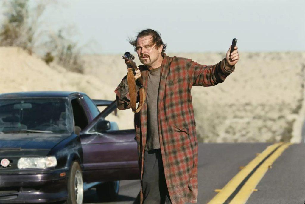Hombre con bigote en camisa a cuadros roja gesticulando junto a auto en carretera desértica