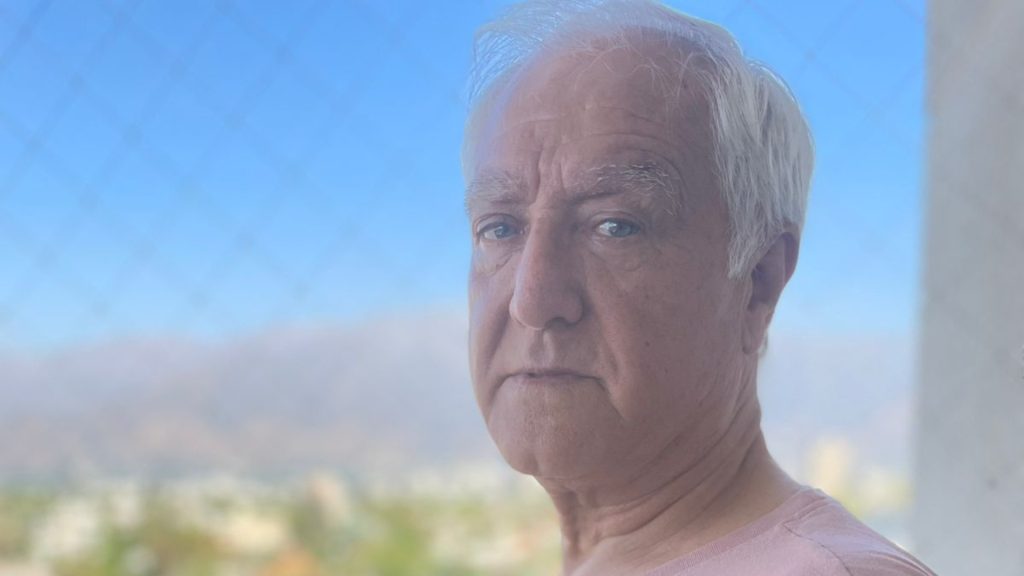 Escritor de mayor edad, cabello blanco, expresión seria, junto a ventana con vista a montañas y cielo despejado