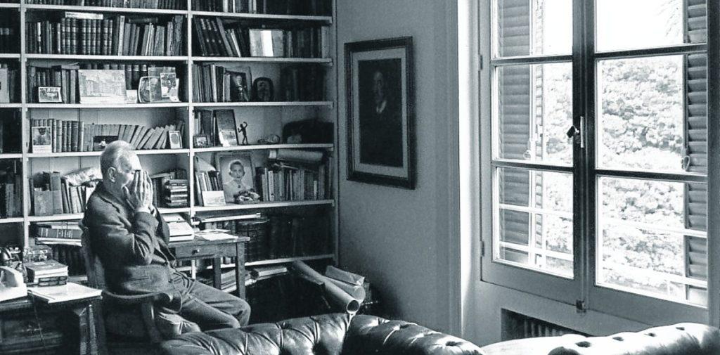 Escritor trabajando en su escritorio frente a libros y ventanas, rodeado de estanterías llenas de libros en blanco