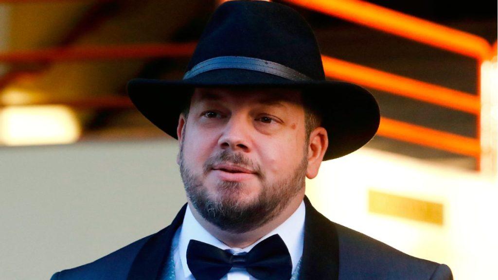 Hombre de traje negro y pajarita blanca con sombrero de fieltro, sobre fondo con luces naranjas