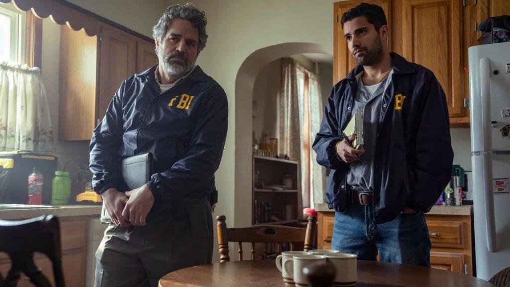 Dos hombres en una cocina desordenada, uno con chaqueta azul sostiene una taza, el otro de perfil también viste