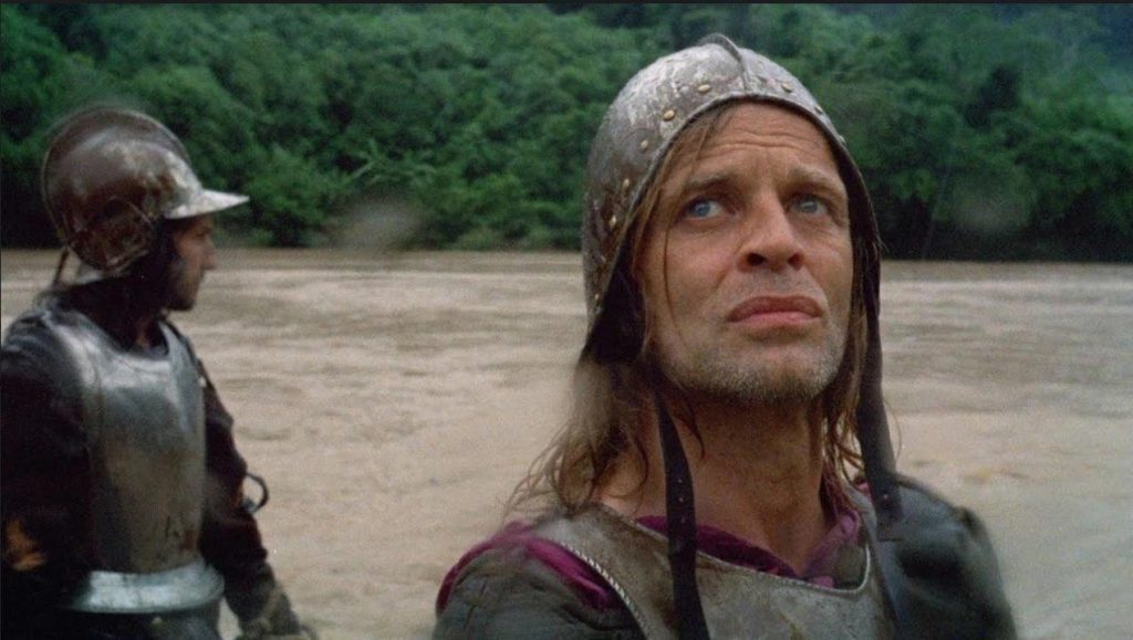 Hombre con cabello largo y ropa medieval junto a río en escena de selva amazónica, expresión de determinación