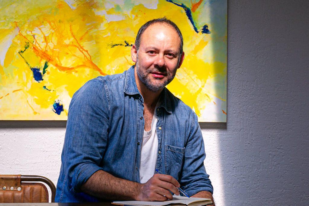 Ignacio Garay sonríe frente a una pared gris con cuadro amarillo de fondo, vistiendo camisa denim azul