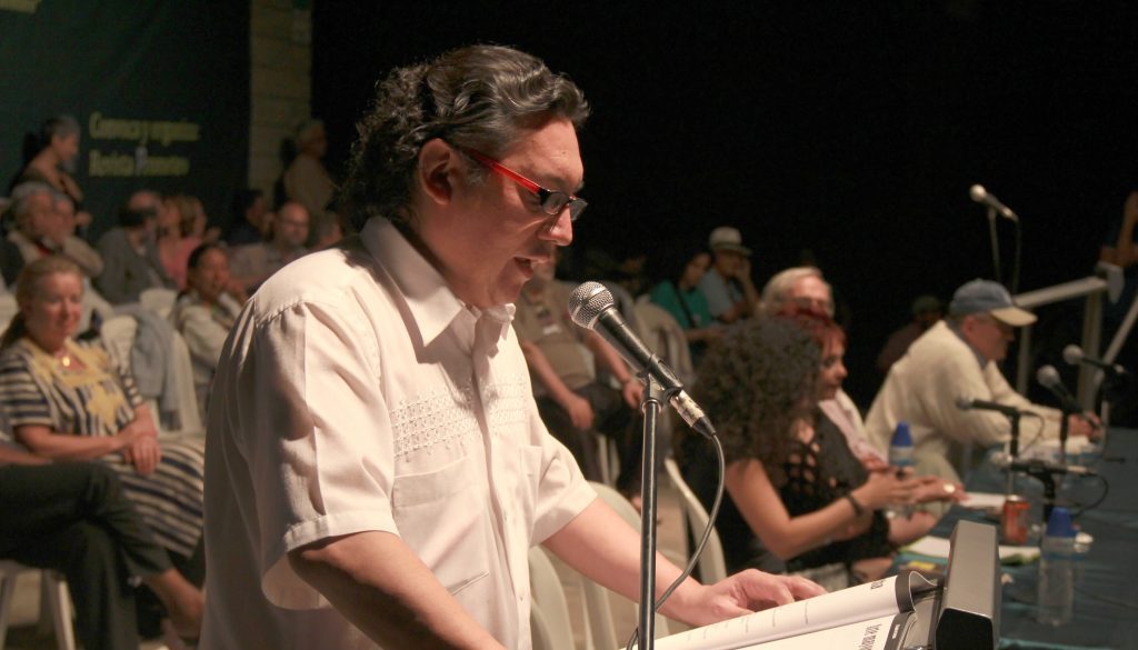 Hombre con gafas en podio con micrófono durante presentación o panel, público al fondo en evento cultural