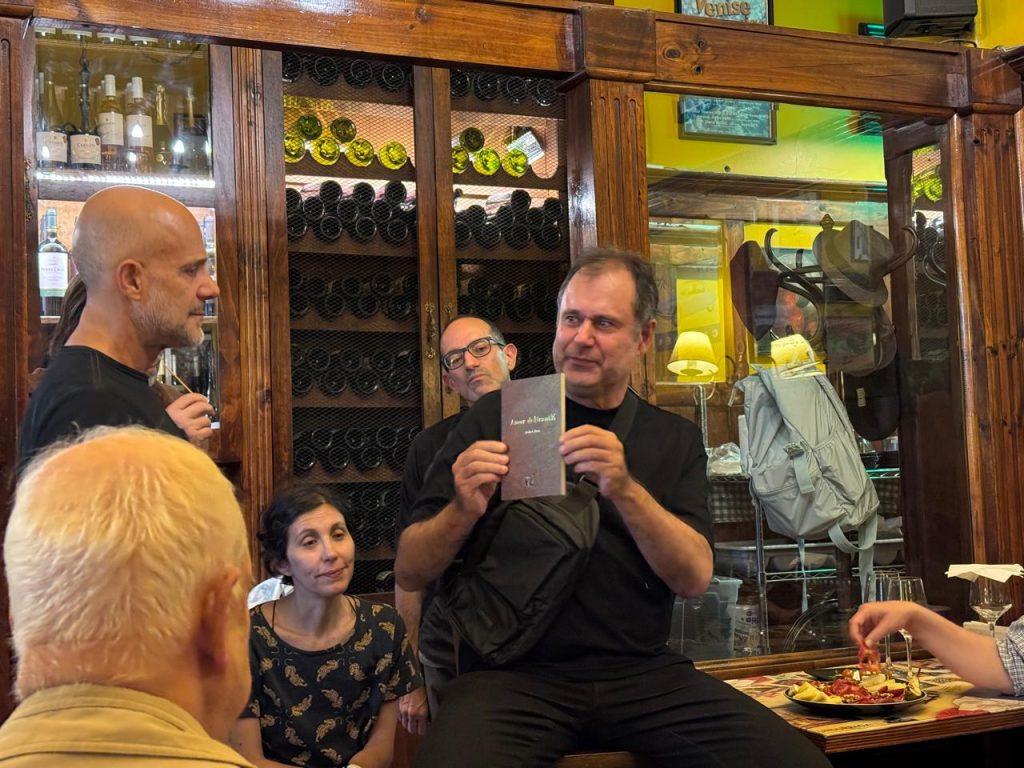 Hombre presentando en biblioteca con botellas de vino y público atento en primer plano, ambiente de evento cultural