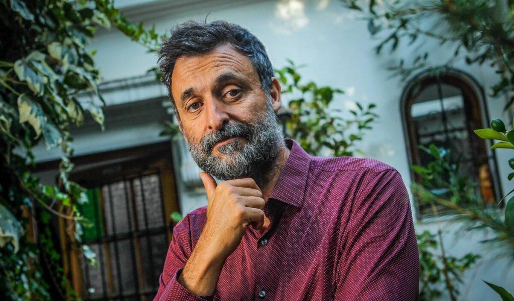 Cristián Warnken, hombre de mediana edad con barba, en pose reflexiva frente a fachada colonial con enredaderas