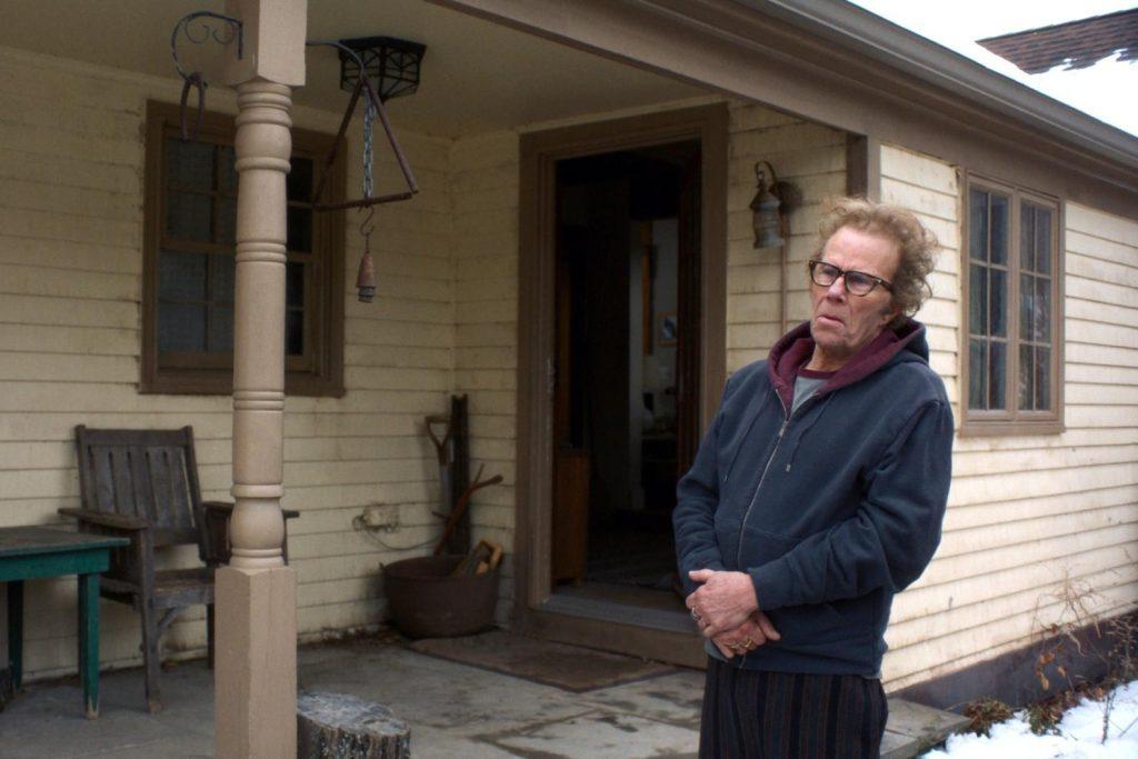 Jim Jarmusch frente a la entrada de una casa de madera blanca, vistiendo chaqueta azul oscuro y gafas