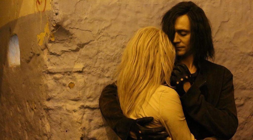 Pareja en escena nocturna de intimidad gótica, él de cabello oscuro y ella rubia, en ambiente de piedra antigua y luz