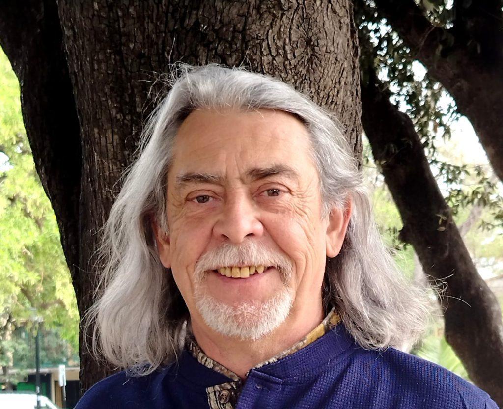 Rubén Torres, escritor chileno de cabello gris largo, sonríe ante un árbol en ambiente natural