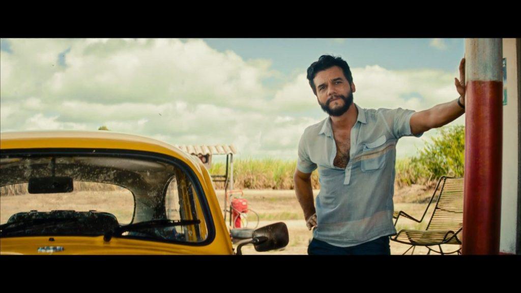 Hombre con barba junto a un automóvil amarillo vintage en una gasolinera desértica, con su brazo extendido
