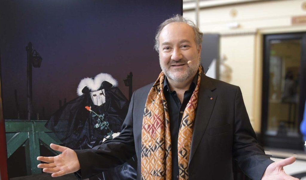 Hombre mayor sonriente junto a póster de película de terror, vistiendo blazer oscuro y bufanda colorida