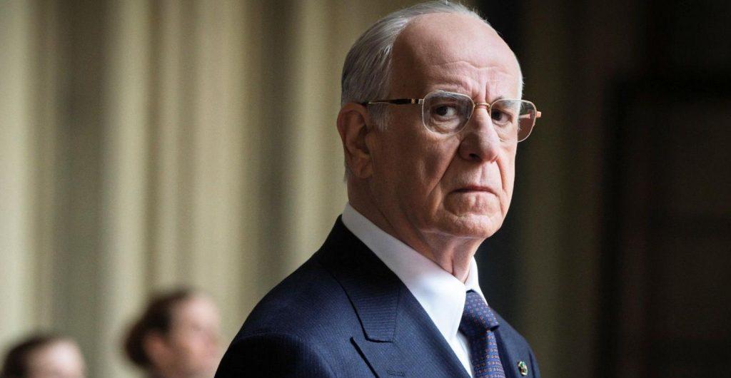 Hombre de edad avanzada con gafas y traje oscuro, expresión seria, en interior formal