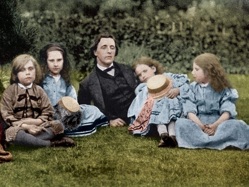 Grupo de cinco niños sentados en el pasto con Lewis Carroll al centro, retrato fotográfico coloreado del siglo XIX