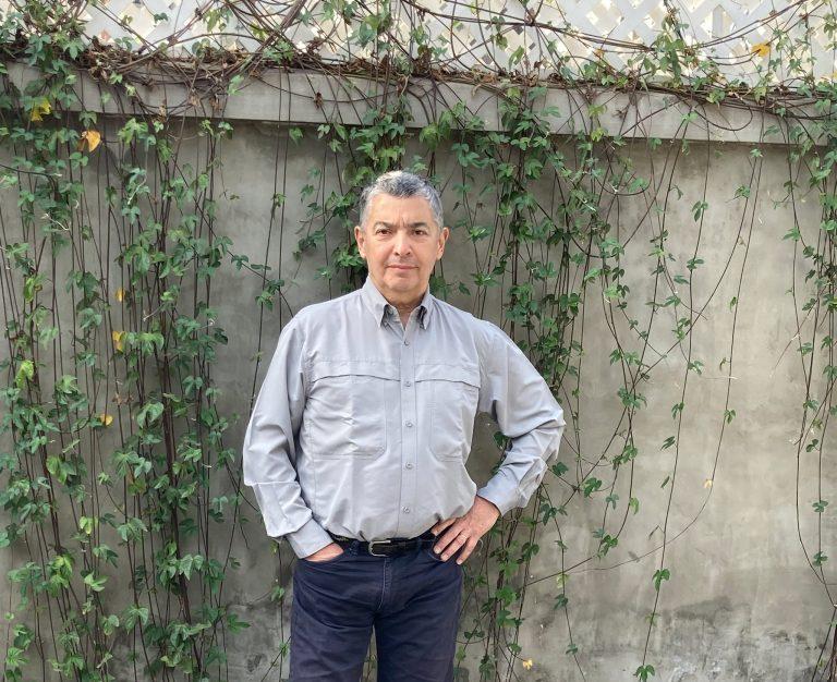 Pablo de Rokha de pie frente a pared de hormigón con enredadera, camisa gris clara y pantalones oscuros