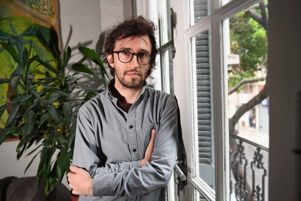 Hombre con gafas y camisa gris de pie junto a una ventana y plantas, posando con los brazos cruzados