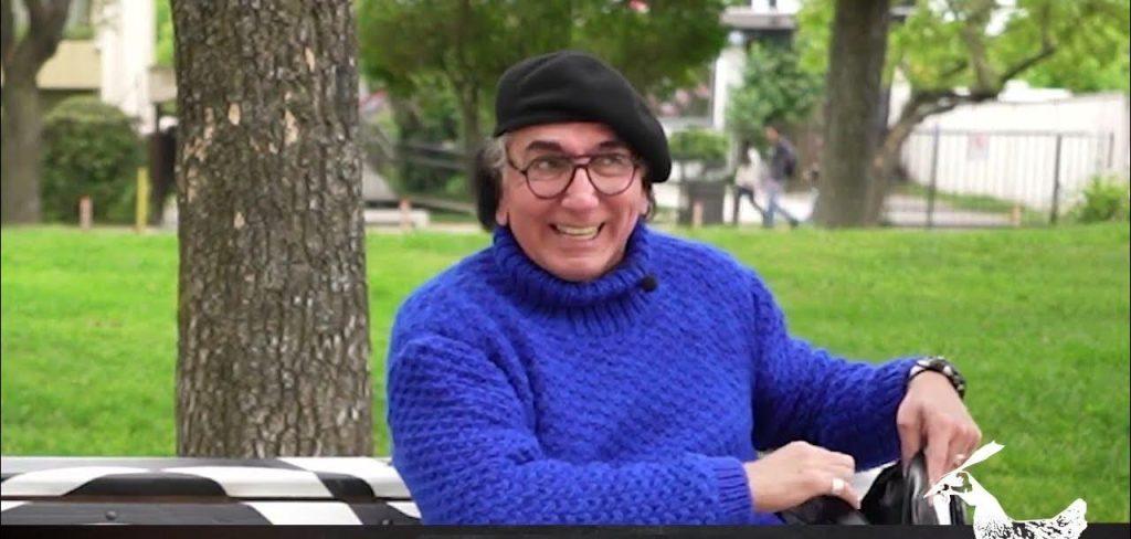 Hugo Godoy, escritor chileno, sentado en un parque bajo un árbol, vistiendo suéter azul y lentes