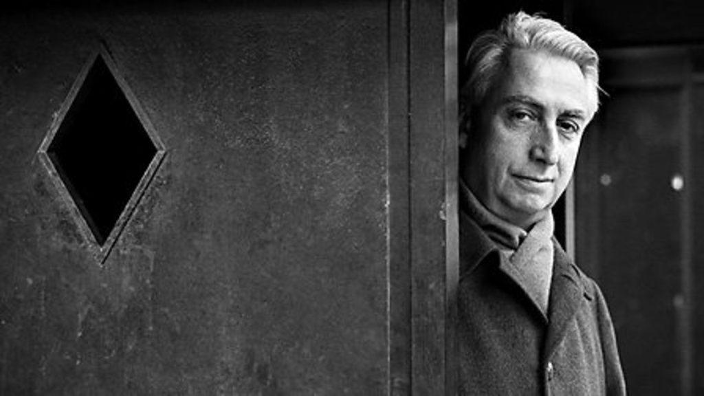 Roland Barthes asomado por una puerta con vidrio de rombo, fotografía en blanco y negro