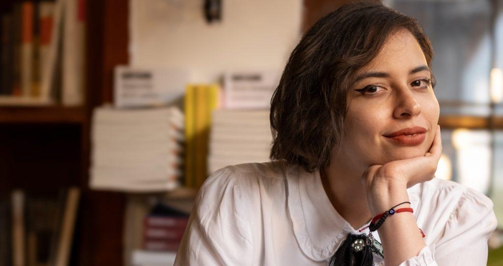 Claudia Amador, escritora, pensativa con la mano en el mentón, en interior con libros de fondo