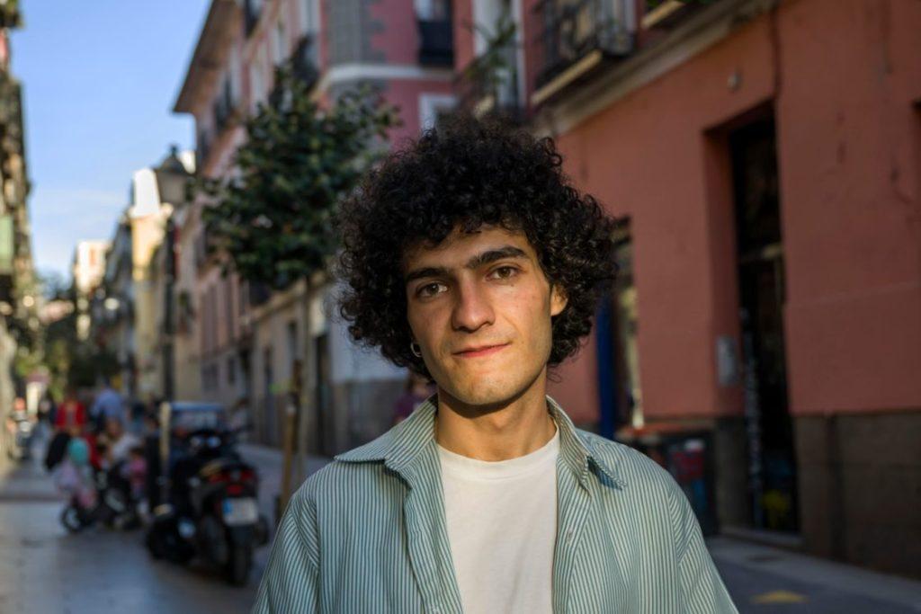 Joven de cabello rizado con camisa gris claro sobre blanca en calle española con edificios antiguos al fondo