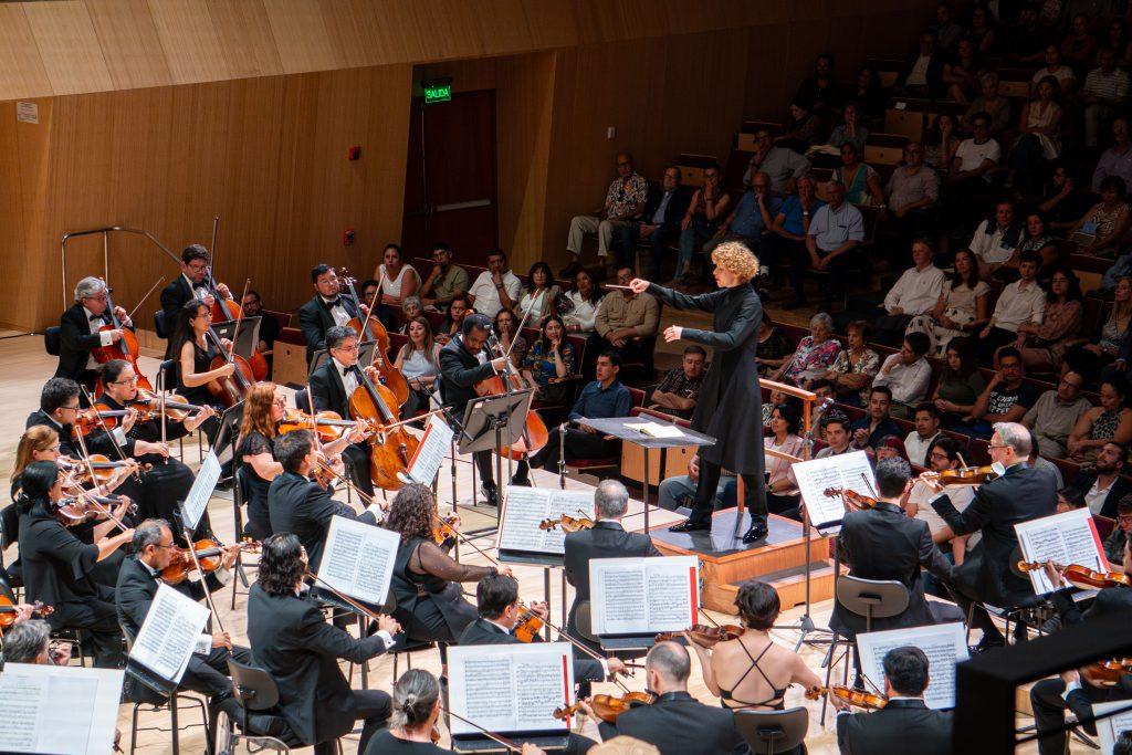 Orquesta sinfónica completa en concierto, músicos con instrumentos de cuerda y viento en la Gran Sala, dirigidos