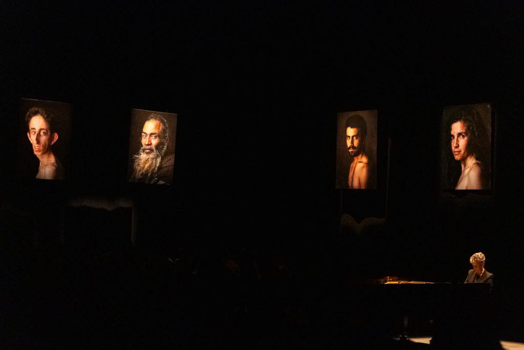 Escena teatral oscura con actores iluminados en varios puntos del escenario, creando una atmósfera dramática