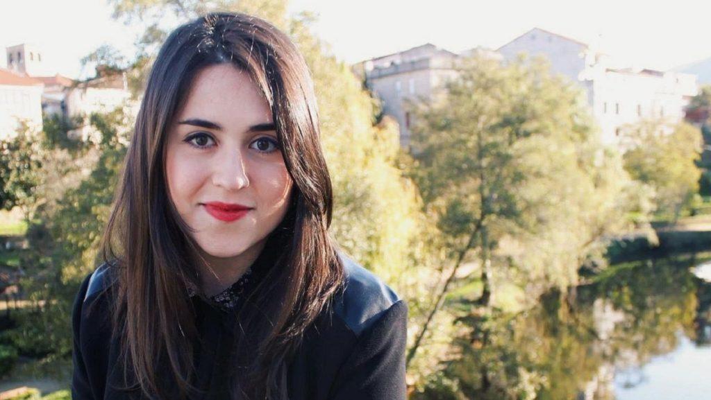 Mujer joven de cabello largo oscuro y labios rojos sonríe a la cámara con un río y árboles desenfocados de fondo