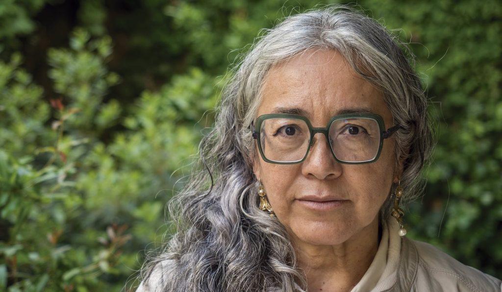 Mujer de cabello gris largo, gafas y expresión serena, con fondo de vegetación verde
