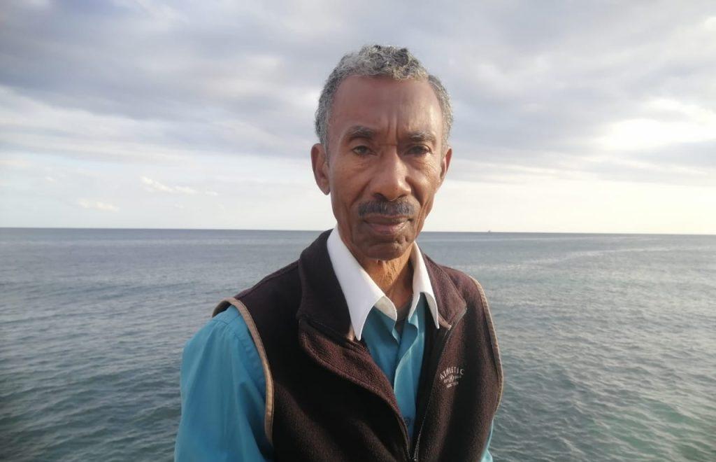 Hombre de mediana edad en primer plano frente al mar, vistiendo chaleco marrón sobre camisa azul, con cielo nublado