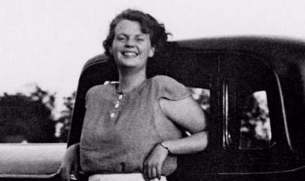 Mujer recostada sobre un automóvil clásico, sonriendo hacia la cámara en fotografía en blanco y negro de época