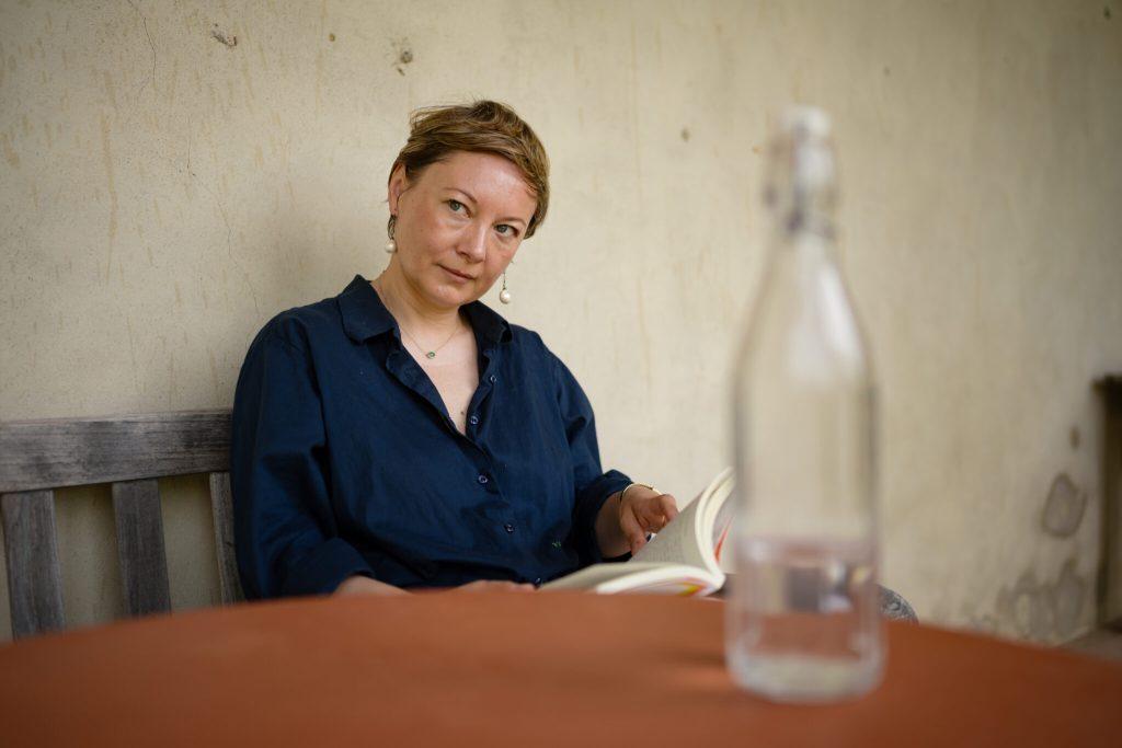 La poeta bielorrusa Valzhyna Mort sentada, con camisa azul, leyendo un libro junto a una mesa con una botella