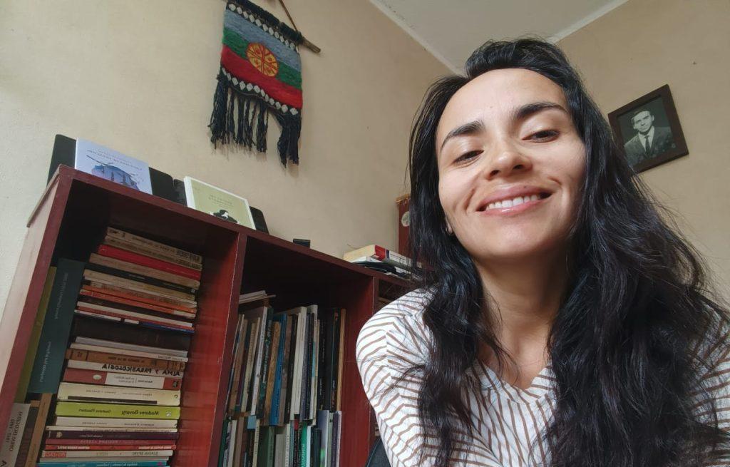 Alejandra Moya Díaz sonríe frente a estantería con libros y objetos decorativos en su hogar