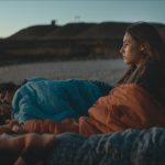 Mujer acostada en la playa al atardecer, vistiendo ropa azul y naranja, con acantilados al fondo